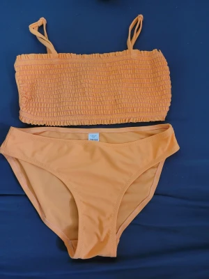 Orange bandeau bikini med smock - Säljer en snygg orange bikini med bandeau-topp och smockdetaljer. Toppen har tunna axelband och bikinitrosorna har klassisk passform. Perfekt för lata dagar på stranden eller poolen.