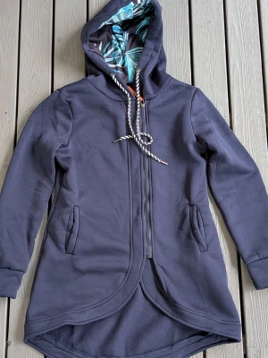 Mörkblå lång hoodie med huva - Snygg mörkblå hoodie med längre passform och rundad nederkant. Utrustad med dragkedja framtill, två fickor och en stor huva med svartvit dragsko. Insidan av huvan har ett mönstrat tyg i blått och vitt. Perfekt för en avslappnad och trendig stil.