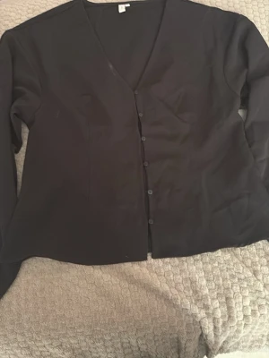 Svart kofta från Nelly, V-ringad - Stilren svart kofta från Nelly med V-ringning och knäppning framtill. Långärmad modell i mjukt tyg som passar perfekt till lager på lager. Enkel och klassisk design som funkar till många olika looks.