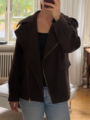 Mörkbrun oversize jacka med dragkedja - Snygg mörkbrun oversize jacka med stor krage och sned dragkedja framtill. Jackan har en loose passform och stora fickor på sidorna. Perfekt för höst och vinter när du vill vara både varm och stilren.