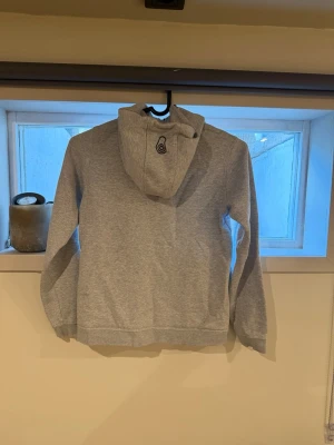 Ljusgrå hoodie med dragkedja - Säljer en ljusgrå hoodie med dragkedja framtill och en bröstficka med dragkedja. Hoodien har en huva med tryckt logga och ribbade muddar vid ärmslut och nederkant. Perfekt för chill dagar eller när du vill ha något bekvämt och stilrent.