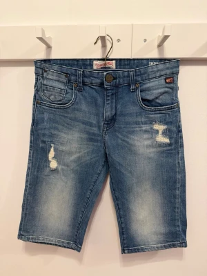 Blå jeansshorts med slitningar - Säljer ett par blå jeansshorts från Petrol Industries med slitna detaljer framtill och klassisk femficksdesign. Shortsen har normal passform, knappgylf och bälteshällor. Perfekta för sommaren och ger en avslappnad streetwear-look. Mycket bra skick, ser ut som nya. Storlek 176/S.