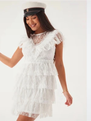 Vit spetsklänning med volanger - Supersöt vit kort spetsklänning med flera lager volanger och v-ringad spets upptill. Klänningen har korta ärmar och en romantisk look som passar perfekt till student eller midsommar. Tillverkad i spets med fina detaljer för en extra festlig känsla. En av öglorna till att knappa där bak har lossnat men syns inte när man har på sig den & fungerar precis som vanligt ändå💌