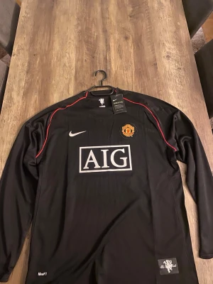 Manchester United långärmad tröja Nike - Snygg svart långärmad Manchester United-tröja från Nike med AIG-logga på bröstet och klubbmärke. Röda detaljer längs axlarna och tillverkad i Dri-FIT material som andas. Perfekt för fotboll eller träning.