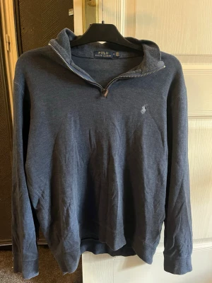 Blå hoodie från Polo Ralph Lauren - Mörkblå hoodie från Polo Ralph Lauren med halv dragkedja och broderad logga på bröstet. Tillverkad i mjukt bomullsmaterial och har ribbade muddar vid ärmslut och nederkant. Perfekt för en avslappnad och stilren look.