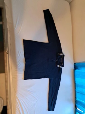 Tommy Hillfiger stickad tröja  - Mörkblå stickad tröja med halv dragkedja vid halsen och ståkrage. Tröjan har långa ärmar och en diskret randig detalj på insidan av kragen. Perfekt för kyliga dagar och enkel att matcha med jeans eller chinos.