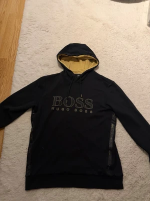 Svart hoodie från Hugo Boss - Svart hoodie från Hugo Boss med stor guldfärgad logga på bröstet och kontrasterande gul insida på huvan. Hoodien har dragsko i huvan och ribbade muddar. Perfekt för en stilren och trendig look.