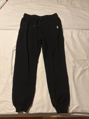 Svarta mjukisbyxor från Polo Ralph Lauren - Svarta mjukisbyxor från Polo Ralph Lauren med klassisk vit broderad logga på vänster sida. Byxorna har dragsko i midjan och muddar vid bensluten. Perfekta för chill dagar eller när du vill vara bekväm. Tillverkade i mjukt material.