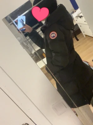 Svart lång dunjacka Canada Goose - Säljer en svart lång dunjacka från Canada Goose med huva och klassisk logotyp på ärmen. Jackan har en figurnära passform, dolda fickor och knappar vid ärmsluten. Perfekt för kalla vinterdagar och riktigt varm. Jackan har fler tecken på användning därav priset. Men knapparna o kedjan funkar som dom ska! Storlek xs