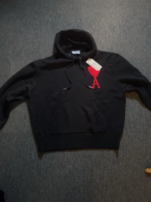 Svart hoodie från Ami med röd logga - Snygg svart hoodie från Ami med stor röd hjärtformad A-logga på bröstet. Klassisk huva med dragsko och magficka framtill. Tillverkad i mjukt material som känns skönt mot huden. Perfekt för en chill och trendig look.