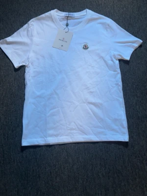 Vit t-shirt från Moncler - Klassisk vit t-shirt från Moncler med rund hals och korta ärmar. T-shirten har Monclers ikoniska logga broderad på bröstet och är tillverkad i mjuk bomull. Perfekt basplagg för en clean och stilren look.
