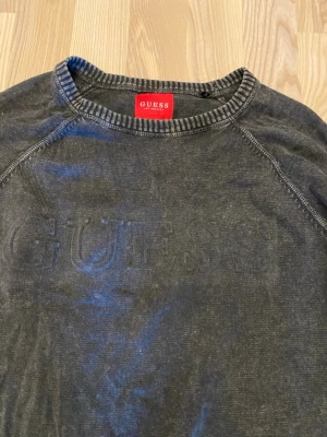 Svart sweatshirt från Guess - Cool svart sweatshirt från Guess med diskret logga broderad på bröstet. Tröjan har rund halsringning, långa ärmar och ribbade muddar vid ärmslut och nederkant. Perfekt till jeans eller joggers för en avslappnad look. Sömmen vid muddarna är något slitna, se bilder. 