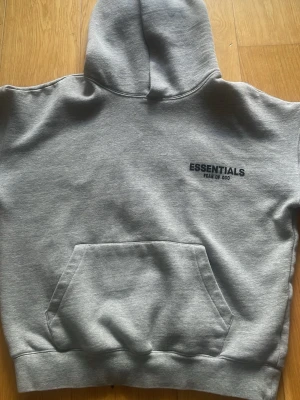Grå hoodie Essentials Fear of God - Säljer en grå hoodie från Essentials Fear of God med svart logga på bröstet. Klassisk känguruficka framtill och stor huva. Tillverkad i mjukt bomullsmaterial som är perfekt enkel och stilren design som passar till allt.