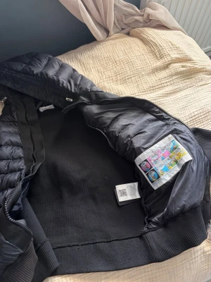 Svart pufferjacka med huva - Snygg svart pufferjacka med huva och ribbade muddar. Jackan har quiltad design, dragkedja framtill och två fickor med knappstängning. Insidan har en färgglad etikett med serietidningsmotiv. Perfekt för kalla vinterdagar.