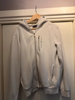 Ljusgrå hoodie med dragkedja - Säljer en vit hoodie med dragkedja framtill och huva. Hoodien har en bröstficka med dragkedja och diskret tryck på bröstet samt ett litet tryck på huvan. Mjukt material och ribbade muddar vid ärmslut och nederkant.