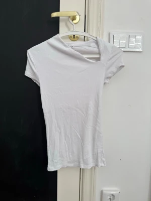 Vit ribbad topp med asymmetrisk hals - Säljer en vit ribbad topp från Zara med korta ärmar och en unik asymmetrisk halsringning. Toppen är figurnära och har en modern, stilren look som passar perfekt till jeans eller kjol.