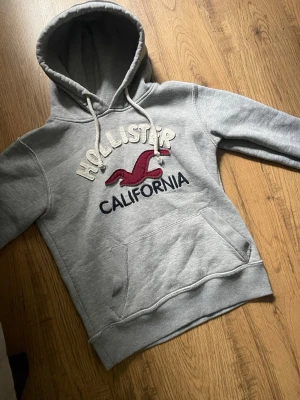 Grå Hollister hoodie med logga - Är medium men har sitter som small. Snygg grå hoodie från Hollister med stor broderad logga och texten 'California' på bröstet. Hoodien har en klassisk känguruficka och justerbar huva med vita snören. Perfekt för en avslappnad och trendig archive stil