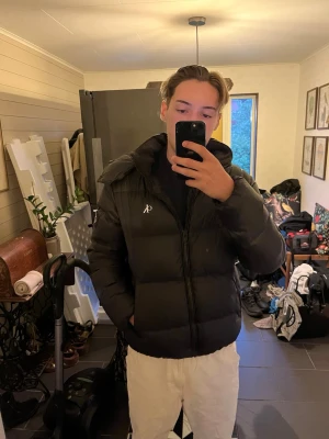 Svart pufferjacka Calvin Klein Jeans - Säljer en svart pufferjacka från Calvin Klein Jeans i storlek M. Jackan har en hög krage, dragkedja framtill och diskret logga på bröstet och armen. Perfekt för kalla vinterdagar och riktigt skön att bära. Klassisk modell som passar till det mesta. 2 små slitningar på bakre vänsterarm som går att sy, en slitning vid högersida luva som också går att fixa, och en liten bit av K;et borta i CK märket på bröstet