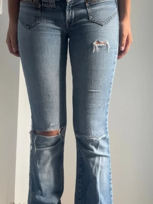 Lågmidjade bootcut jeans vintage - Säljer ett par ljusblå bootcut jeans med slitningar och hål vid knäna och låret. Jeansen har dragkedjedetaljer vid fickorna och klassisk femficksdesign. Tillverkade i denim med en cool vintagekänsla. Insydda vid midjan och uppkortade i benen passar i längd för mig (166cm). Motsvarar XS i strl. 