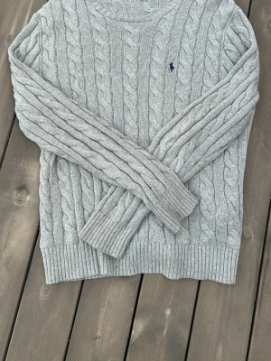 Grå kabelstickad tröja Polo Ralph Lauren - Snygg grå kabelstickad tröja från Polo Ralph Lauren med rund halsringning och diskret broderad logga på bröstet. Tröjan har långa ärmar och ribbade muddar vid ärmslut och nederkant. Perfekt för lager på lager och klassisk stil.