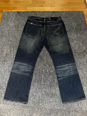 Mörkblå Ed Hardy jeans med brodyr - Om det undras om mått så är de bara fråga så löser jag! Säljer ett par mörkblå jeans från Ed Hardy med raka ben och snygga slitningar. Bakfickan har broderad Ed Hardy-logga i vitt och silver. Jeansen har klassisk femficksmodell och kontrastsömmar. Perfekta för dig som gillar streetwear och detaljer.