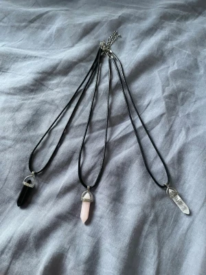 Halsband med kristallhänge -  En för 40 och tre för 100.Tre halsband med svarta band och spetsiga kristallhängen i svart, rosa och transparent. Hänget sitter i ett silverfärgat fäste och ger en stilren och trendig look. Perfekt accessoar för dig som gillar kristaller och minimalistisk stil.