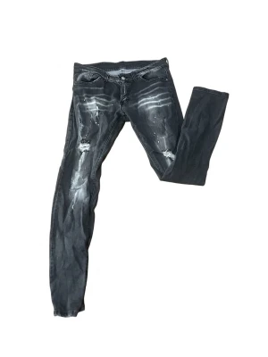 Svarta DSQUARED2 skinny jeans med slitningar - Säljer ett par svarta skinny jeans med coola slitningar och vita färgstänk. Jeansen har fem fickor, låg midja och detaljerade slitningar både fram och bak. Perfekta för dig som gillar en edgy streetstil.