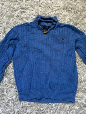 Blå kabelstickad tröja med halvzip - Snygg blå kabelstickad tröja med halvzip och ståkrage. Tröjan har långa ärmar, ribbade muddar och en liten broderad logga på bröstet. Nypris 1795kr. I barnstorlek ”XL” motsvarar s/m (163-174cm). Unisex