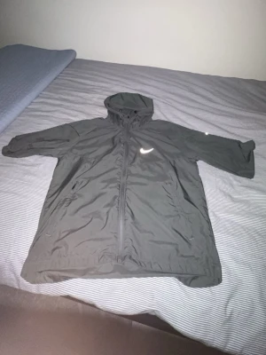Nike Miller jacka  - Snygg svart vindjacka från Nike med dragkedja framtill och praktisk huva. Jackan har två sidofickor och reflekterande Nike-logga på bröstet. Perfekt för träning utomhus eller som ett extra lager under kyliga dagar.