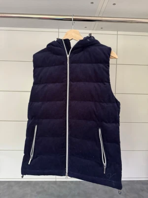 Mörkblå vadderad dunväst med huva - Snygg mörkblå dunväst med huva och dragkedja framtill. Västen har två framfickor med dragkedjor och quiltad design. Materialet ser ut att vara mjukt och lite glansigt, perfekt för lager-på-lager under kyliga dagar.