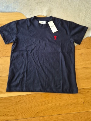 Mörkblå t-shirt från Ami - Snygg mörkblå t-shirt från Ami Alexandre Mattiussi med klassisk passform. T-shirten har en liten röd broderad logga på bröstet och är gjord i mjuk bomull. Perfekt till jeans eller shorts för en clean look.