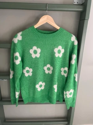 Grön stickad tröja med blommor Lindex - Grön stickad tröja från Lindex med vita blommor över hela plagget. Tröjan har rund halsringning, ribbade muddar och en avslappnad passform. Perfekt för dig som gillar färg och mönster!