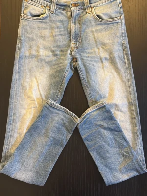 Ljusblå raka jeans med slitningar - Säljer ett par ljusblå jeans ifrån Nudie som har modellen ”Lean Dean” Jeansen har klassisk femficksdesign, kontrastsömmar och detaljerade bakfickor med broderi. Perfekta för en avslappnad och trendig look. Liten defekt som är med på bild 3 som inte märks av mycket vid användning.