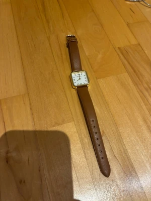 Klocka med brunt läderband - Stilren klocka med fyrkantig urtavla i guldton och vitt. Klockan har ett klassiskt brunt läderband och tydliga svarta siffror på urtavlan. Perfekt accessoar för dig som gillar enkel och tidlös design.