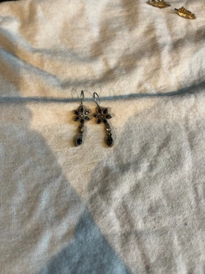 Silvriga blommiga hängörhängen - Säljer ett par silvriga örhängen med blomformad design och små svarta stenar. Örhängena har en hängande detalj under blomman och är tillverkade i metall. Perfekta för dig som gillar unika och detaljerade smycken.