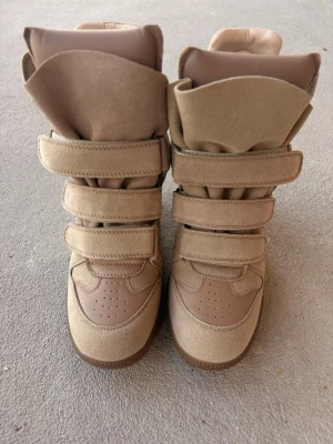 Beige höga sneakers med kardborre - Snygga beige sneakers med högt skaft och tre breda kardborreband. Tillverkade i mockaliknande material med detaljer i syntet. Tjock sula i gummi och vadderad plös för extra komfort. Perfekta för dig som gillar streetstyle och vill sticka ut.