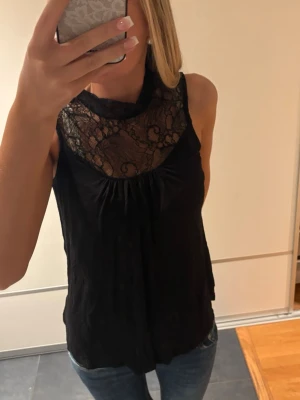Svart ärmlös topp med spetsdetalj - Snygg svart ärmlös topp med transparent spets upptill och rund halsringning. Toppen har en lös passform och är perfekt att styla med jeans eller kjol. Spetsen ger en elegant touch och materialet känns lätt och bekvämt.