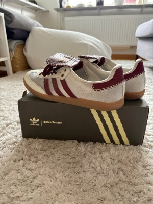 Adidas x Wales Bonner sneakers vinröd/vit - Unika sneakers från Adidas x Wales Bonner i vitt och vinrött med klassiska tre ränder på sidan. Skorna har en cool detalj med kontrastsömmar och bred plös med logga. Ovandelen är i mocka och läder, och sulan är i gummi. Perfekta för dig som vill sticka ut.