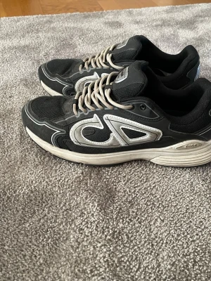 Svarta sneakers med vita detaljer - Säljer ett par svarta sneakers med vita och grå detaljer samt tjock vit sula. Skorna har en sportig look med CD-logga på sidan och bak. Ovandelen är i mesh och syntetmaterial, och de har vita snören. Perfekta för dig som gillar streetwear och chunky sneakers. Står 44 men Passar 41 42 en sko har en annan sula däravpriset 