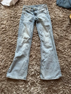 Ljusblå bootcut jeans med slitningar - Säljer ett par ljusblå bootcut jeans med lätt slitna detaljer och klassisk femficksmodell. Jeansen har vida ben och en grön patch bak i midjan. Perfekta för dig som gillar retrostil och vill ha ett par riktigt snygga jeans i garderoben.