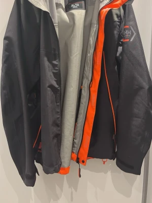 Svart vindjacka med orange detaljer XL - Snygg svart vindjacka från Helly Hansen med grått innerfoder och orange detaljer längs dragkedjan. Jackan har huva, justerbara muddar och logga på ärmen. Perfekt för blåsiga dagar och outdoor-aktiviteter.