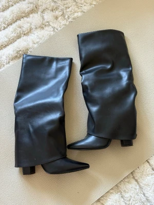Svarta stövlar - Säljer ett par svarta overknee stövlar i slätt, glansigt material med bred klack och spetsig tå. Stövlarna har en loose fit runt skaftet och en dragkedja. Knappt använda