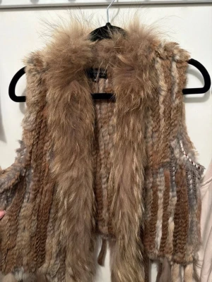 Brun fluffig pälsväst - Säljer en supermysig pälsväst i olika bruna och beige toner. Västen har en fluffig krage och är gjord av mjukt, långhårigt fuskpälsmaterial som ger en riktigt lyxig känsla. Perfekt för att lyfta vilken outfit som helst med lite extra edge.