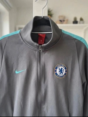 Chelsea Nike grå träningsjacka - Snygg grå träningsjacka från Nike med Chelsea FC-logga på bröstet och ljusblå detaljer på axlarna. Jackan har hel dragkedja, ståkrage och två sidofickor. Perfekt för fotbollsträning eller som sportig vardagsstil.