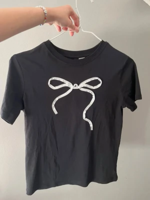 Svart t-shirt med vit rosett - Snygg svart t-shirt med korta ärmar och en vit rosett tryckt på bröstet. Aldrig använd. Hör av er vid frågor!