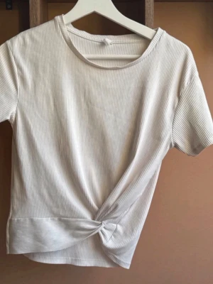 Vit ribbad t-shirt med knutdetalj - Säljer en stilren vit t-shirt med ribbad struktur och en snygg knutdetalj framtill. T-shirten har korta ärmar och rund halsringning. Perfekt för dig som gillar enkla men trendiga plagg.