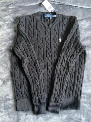 Mörkgrå kabelstickad tröja Polo Ralph Lauren - Snygg mörkgrå kabelstickad tröja från Polo Ralph Lauren med klassisk rund halsringning och vit broderad logga på bröstet. Tröjan har ribbade muddar vid ärmslut och nederkant och är tillverkad i mjukt material. Perfekt för dig som gillar stilrena plagg.