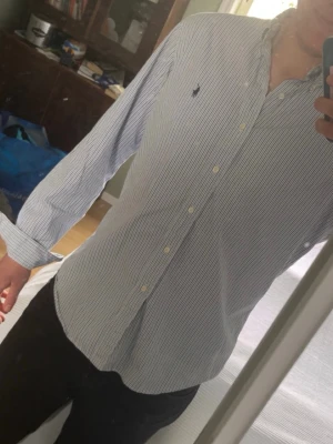 Ralph Lauren skjorta  - Snygg blårandig skjorta med klassisk krage och knäppning framtill. Skjortan har långa ärmar och en liten broderad logga på bröstet. Perfekt till jeans eller chinos för en clean look.
