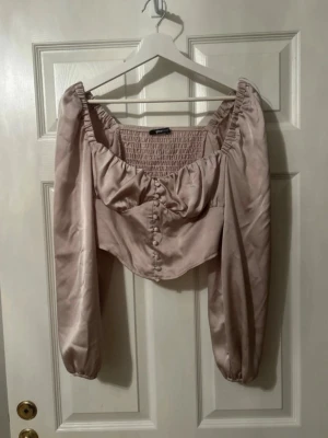 Beige satintopp Gina Tricot - Supersöt beige topp från Gina Tricot i satinliknande material. Toppen har långa puffärmar, knappar framtill och smock i ryggen. Croppad modell med romantisk känsla och lätt glansig finish. Perfekt för dig som vill sticka ut lite extra.