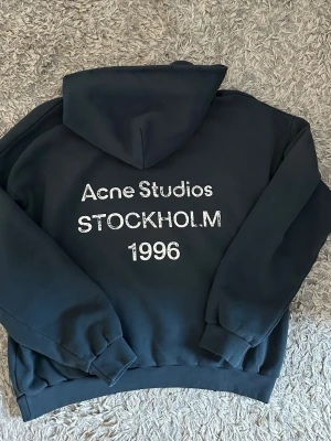 Acne Studios hoodie - Hoodie från Acne Studios med stor huva och tryck på ryggen: 'Acne Studios STOCKHOLM 1996' i vitt. Oversized passform med ribbade muddar vid ärmslut och midja. Öppen för bud!